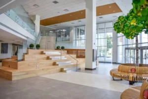 Modern lobby featuring wooden steps, glass railing, communal seating, and greenery, illuminated by natural light and minimalist decor.