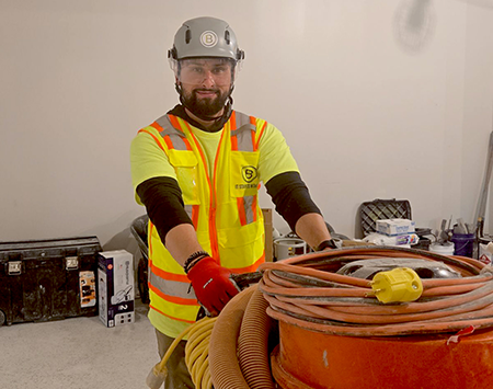 Construction worker in high-visibility vest and helmet handling large orange industrial vacuum with coiled hoses indoors.