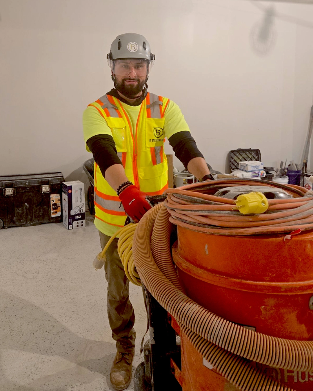 Construction worker in high-visibility vest and helmet handling large orange industrial vacuum with coiled hoses indoors.