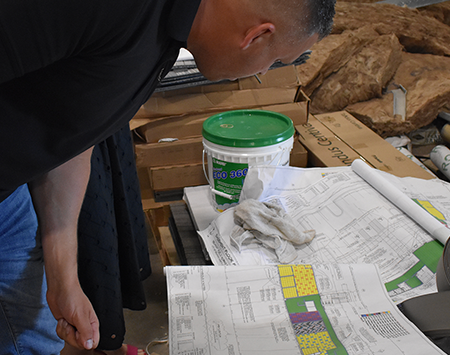 Man in black shirt and jeans leans over a table with building blueprints, a green bucket, and construction materials nearby.