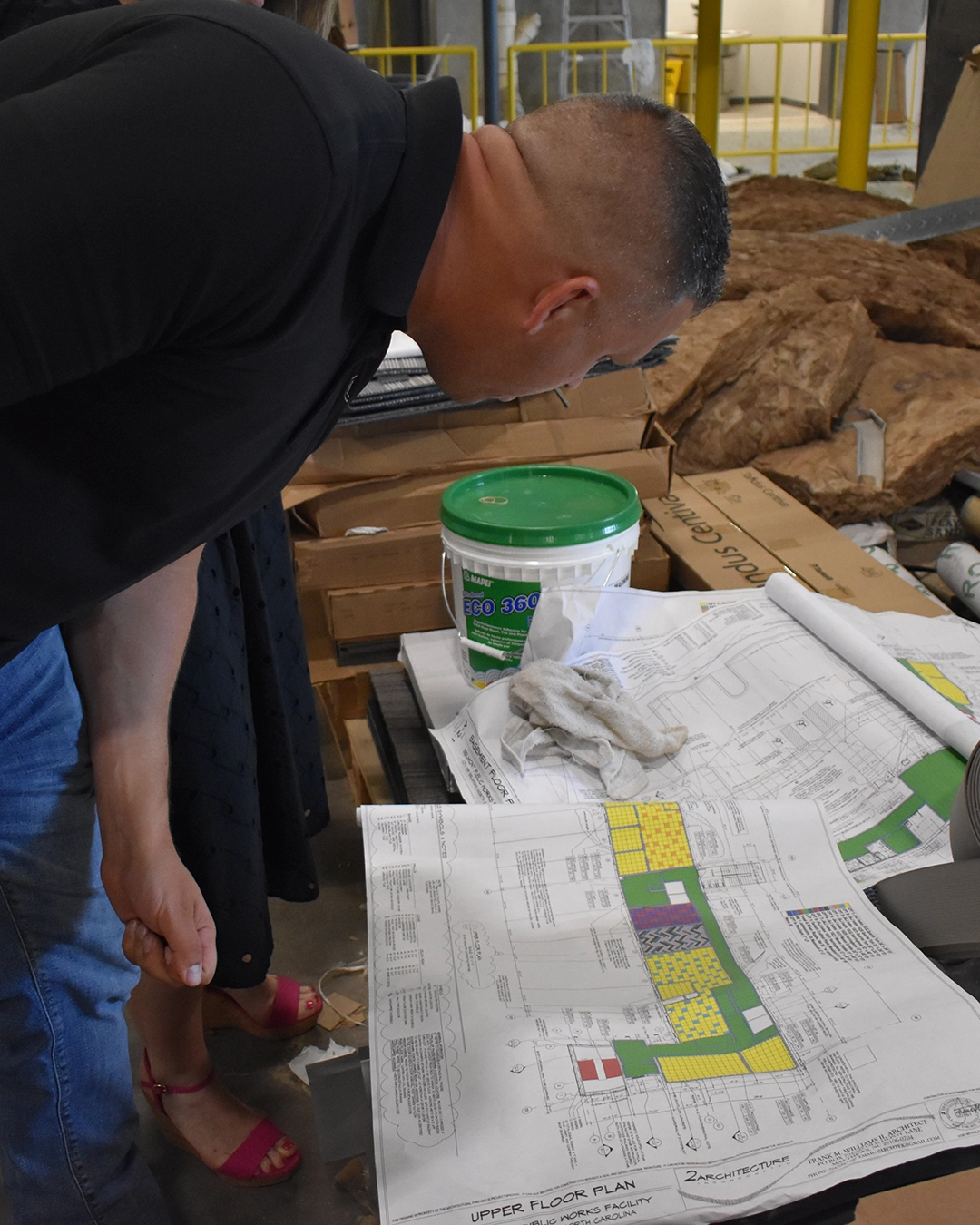 Man in black shirt and jeans leans over a table with building blueprints, a green bucket, and construction materials nearby.