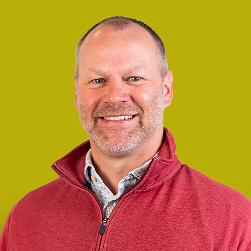 Smiling middle-aged man with short hair and beard wearing a red zip-up sweater and patterned shirt, posed against a yellow background.