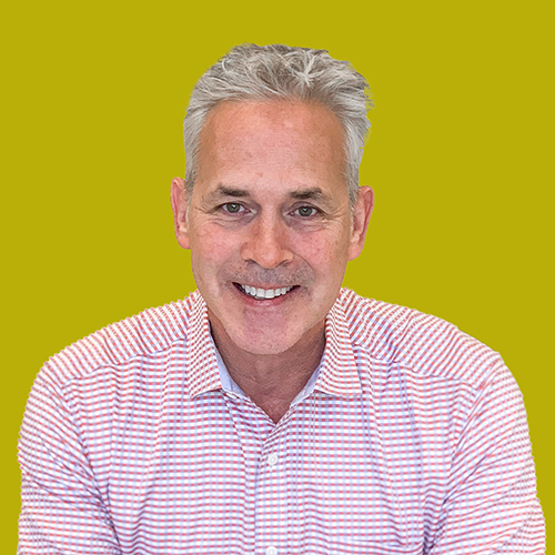 Smiling man with gray hair in a red and white striped shirt sits in front of a plain yellow background.