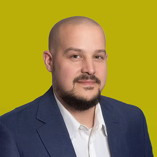 A man with a shaved head, trimmed beard, and mustache, wearing a blue suit jacket and white shirt, poses against a solid yellow background.