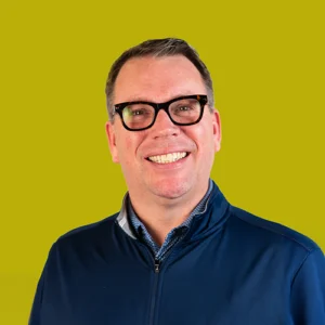 Smiling man with short brown hair and glasses wearing a navy zip-up jacket, posed in front of a solid yellow-green background.