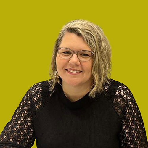 A woman with wavy blonde hair and glasses, wearing a black top with lace sleeves, sits and smiles in front of a yellow-green background.