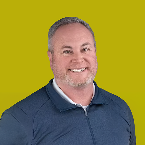 Smiling man with short gray hair and beard wearing a dark blue zip-up jacket over a white shirt, posing in front of a solid yellow-green background.