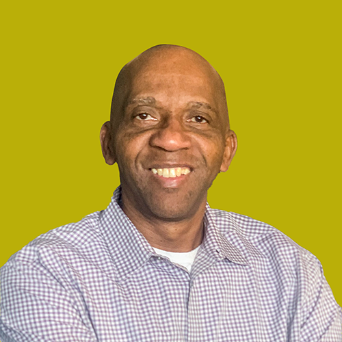 A man wearing a checked shirt smiles at the camera in front of a plain yellow-green background.