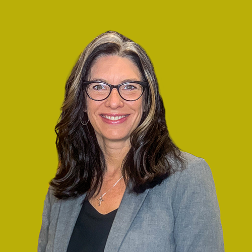 A woman with long dark hair and glasses smiles while wearing a gray blazer over a black top, standing in front of a plain yellow background.