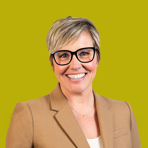 Smiling person with short blonde hair and black glasses wearing a tan blazer, standing in front of a solid yellow background.