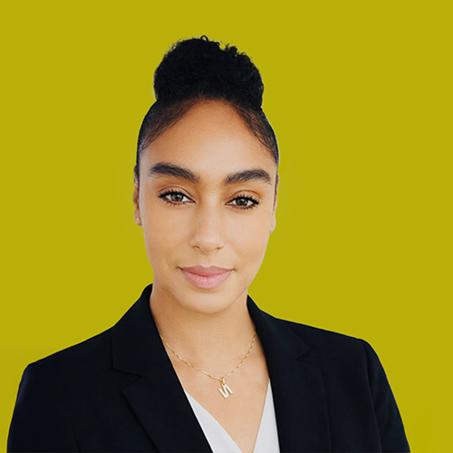 Woman with dark hair in a bun, wearing a black blazer and white top, faces the camera against a solid yellow-green background.