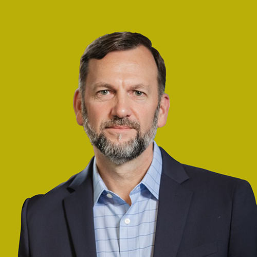 A man with short brown hair and a beard, wearing a blue suit jacket and light blue checkered shirt, stands in front of a plain yellow background.