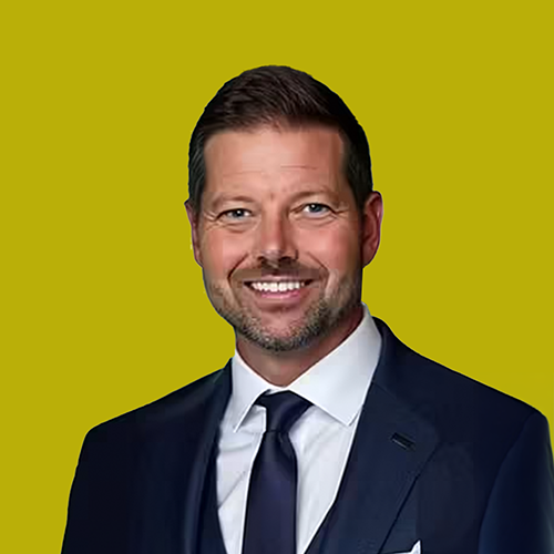 A man in a dark suit, white shirt, and tie smiles at the camera against a solid yellow background.
