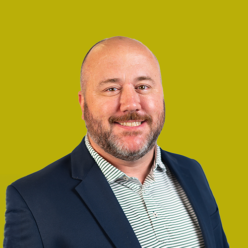 Bald man with a beard wearing a navy blazer and striped shirt, smiling in front of a solid yellow-green background.