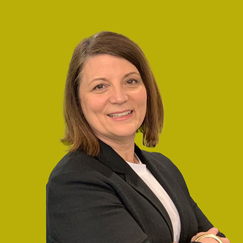 A woman with straight brown hair and a black blazer smiles with folded arms against a solid yellow-green background.