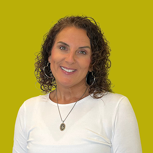 Woman with curly brown hair, wearing a white shirt and necklace, smiles at the camera against a solid yellow background.