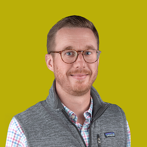 Man with short hair, glasses, and beard wearing a gray vest and plaid shirt, smiling in front of a yellow background.