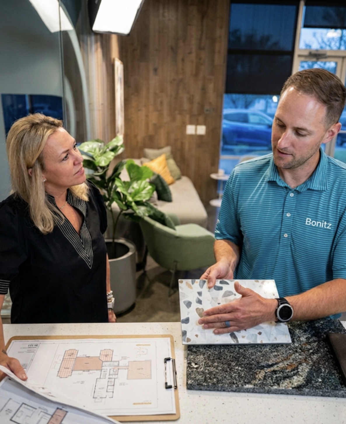 A man shows a woman a terrazzo tile sample while discussing floor plans on a clipboard in a modern office setting.