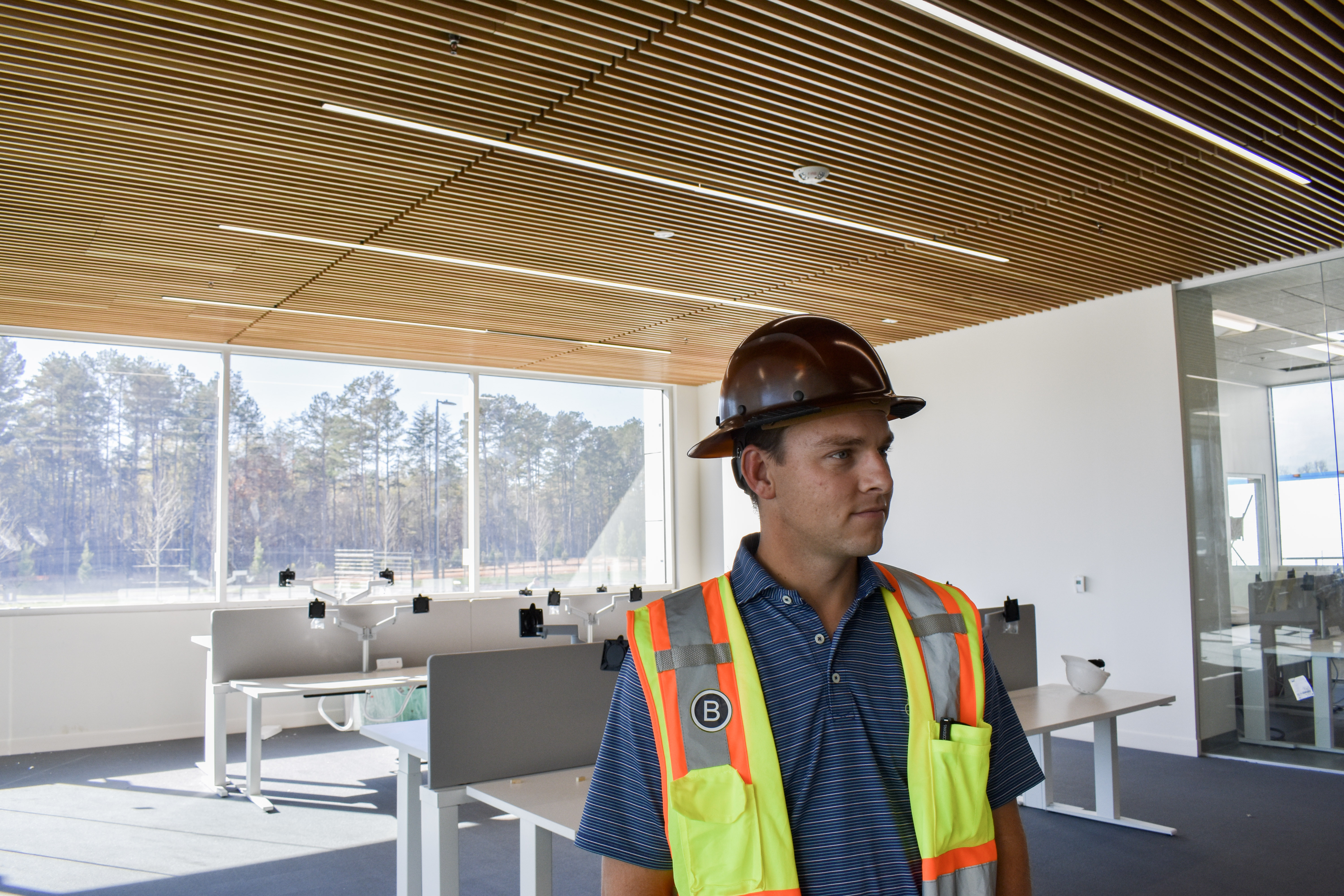 A man in a hard hat and safety vest stands in a modern, partially furnished office space with large windows and wooden ceiling panels.