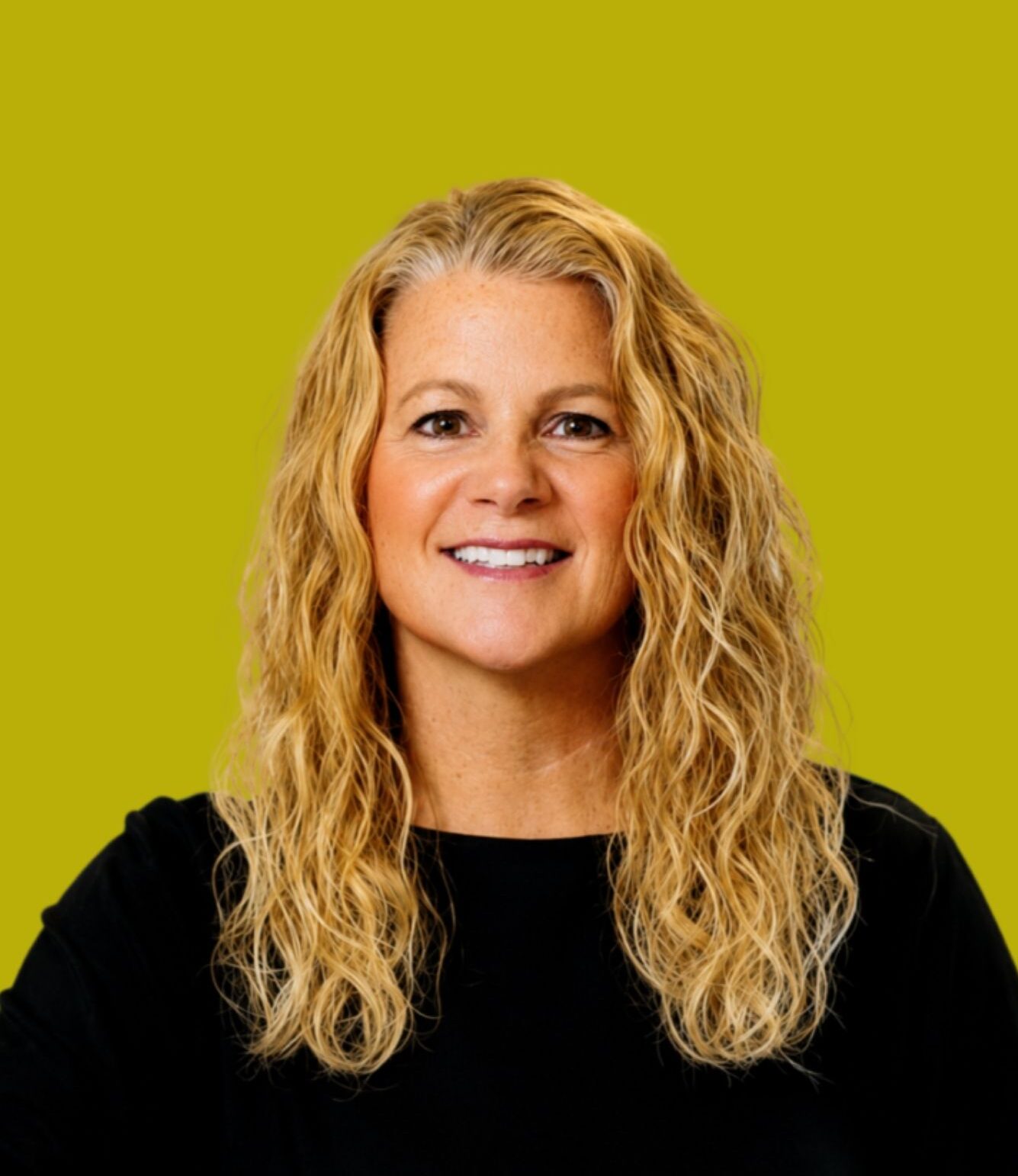 Smiling woman with wavy blonde hair wearing a black top, posed in front of a solid yellow-green background.