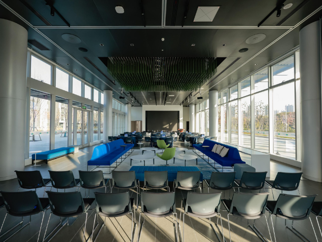 Modern conference room with rows of black chairs, blue couches, a green chair, large windows, overhead lighting, acoustical solutions for sound clarity, and a screen at the front.