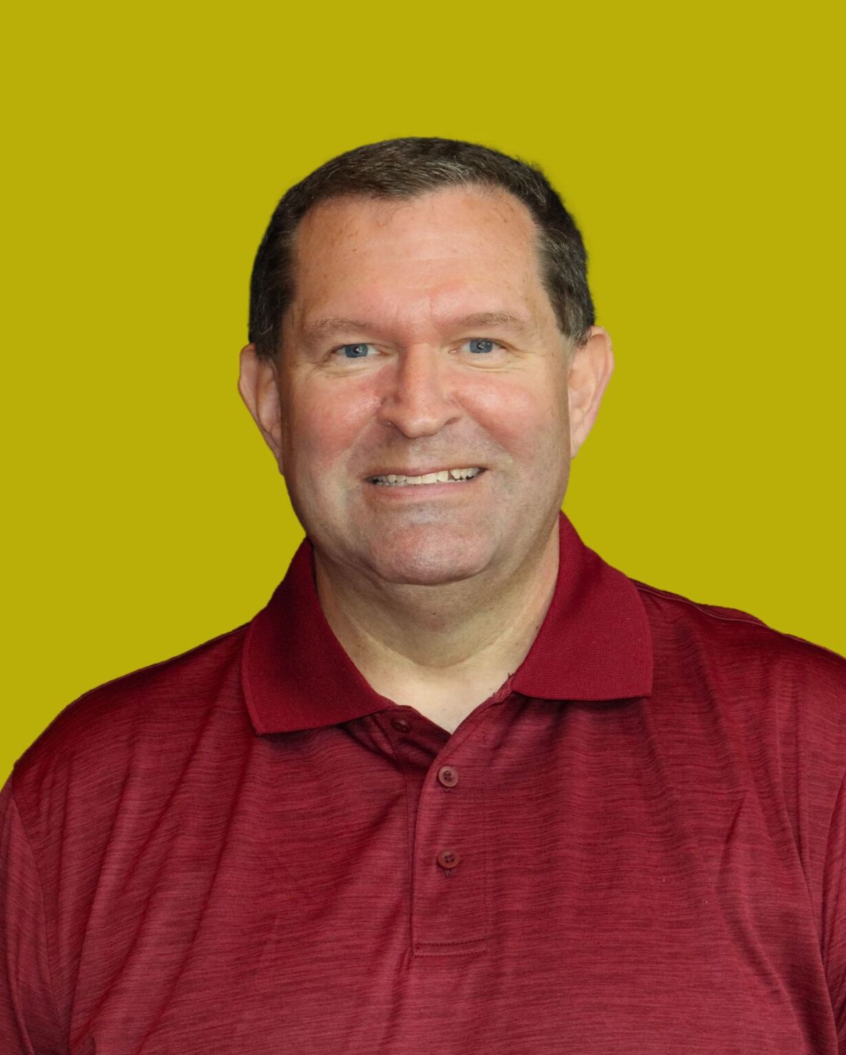 A man with short brown hair, wearing a maroon polo shirt, smiles at the camera against a solid yellow background.