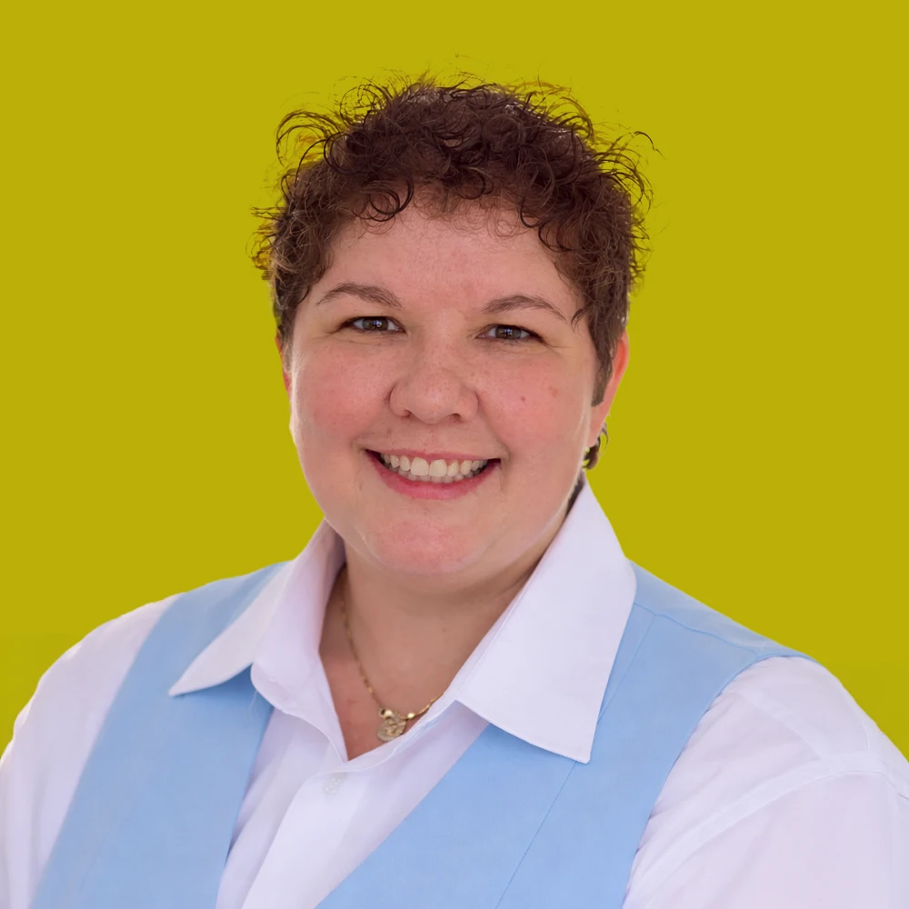 Smiling person with short curly hair wearing a white shirt and light blue vest, posed against a solid yellow background.