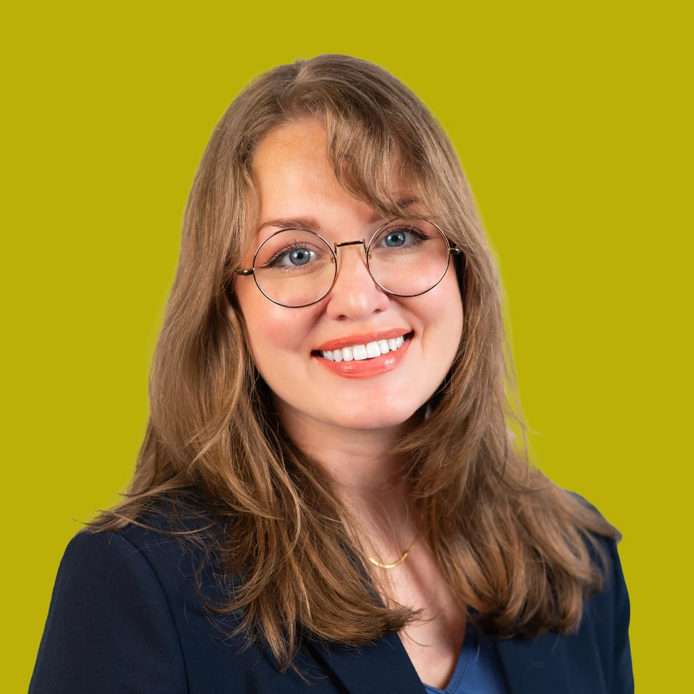 Smiling woman with long light brown hair and glasses, wearing a dark blazer and blue top, posed against a yellow-green background.