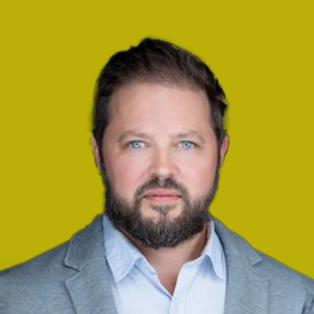 A man with a beard and short hair, wearing a light gray blazer over a white shirt, stands against a plain yellow background.