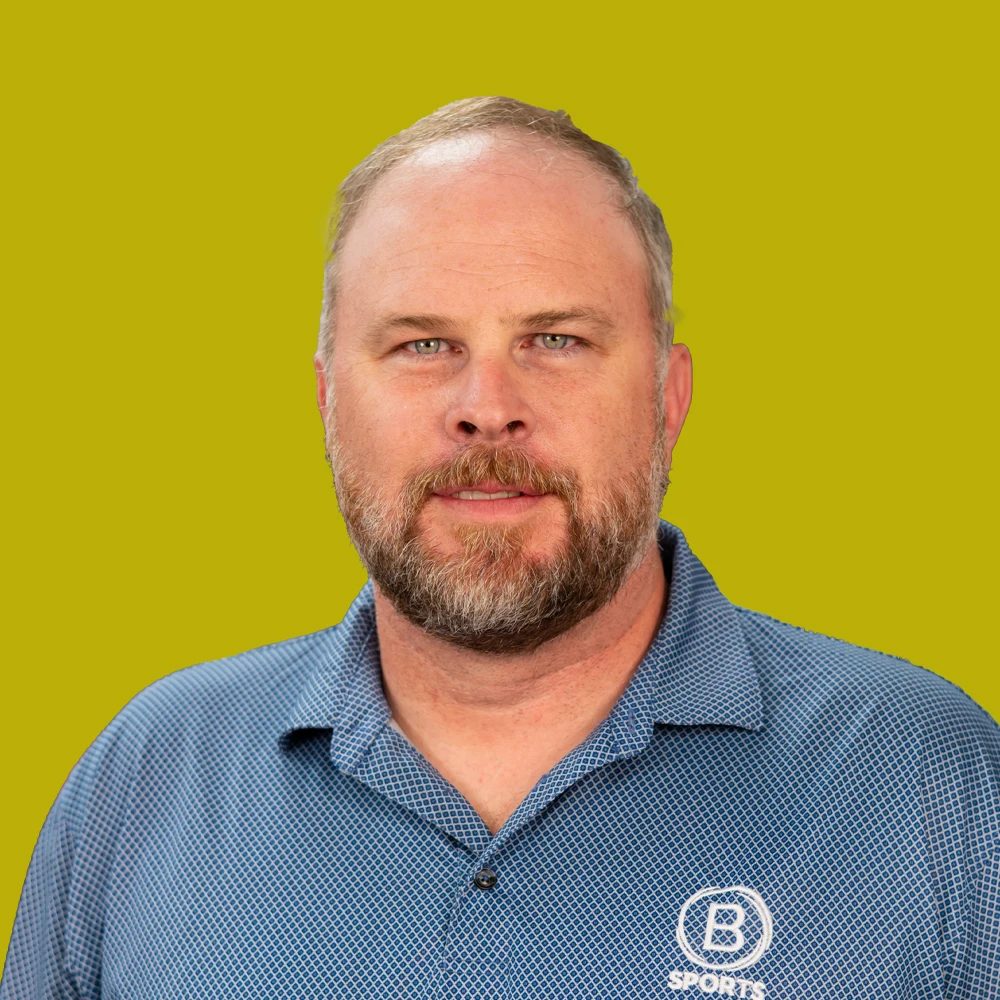 A man with a short beard and mustache wears a blue collared shirt and stands against a plain yellow background.