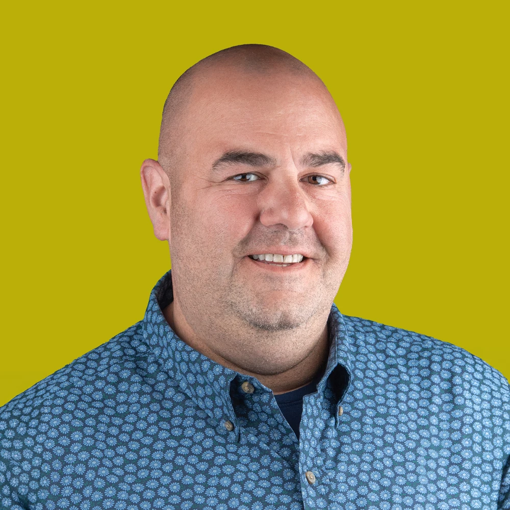 A man with a shaved head and patterned blue shirt smiles at the camera against a solid yellow-green background.