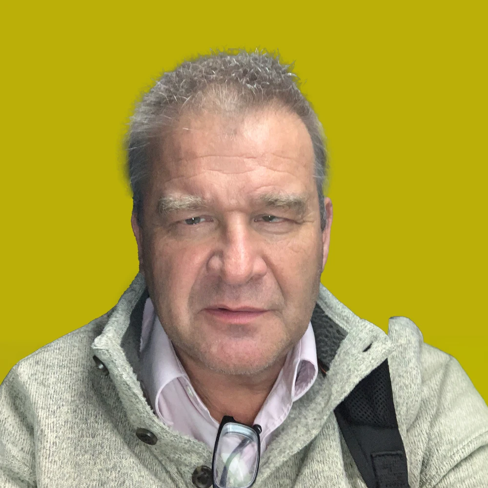 Middle-aged man with short gray hair wearing a light gray sweater and pink shirt, sitting against a solid yellow background. Glasses rest on the front of his sweater.