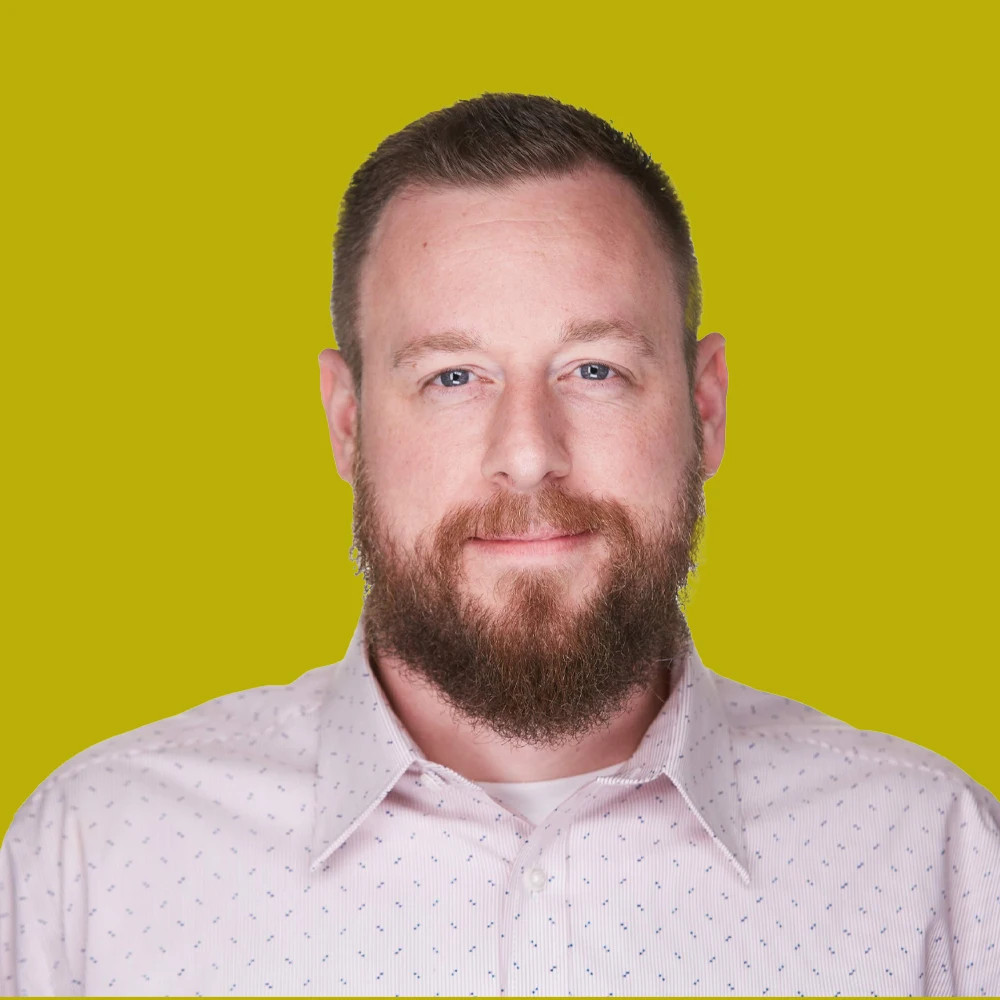 A man with a brown beard and short hair, wearing a light pink button-up shirt, stands in front of a solid yellow-green background.