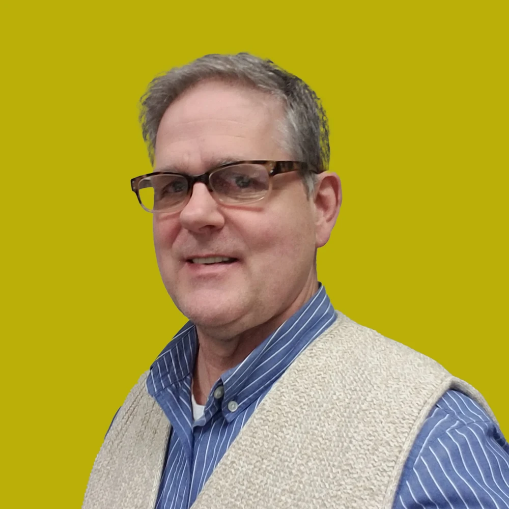 Middle-aged man with short gray hair and glasses, wearing a blue striped shirt and beige vest, smiling in front of a solid yellow background.