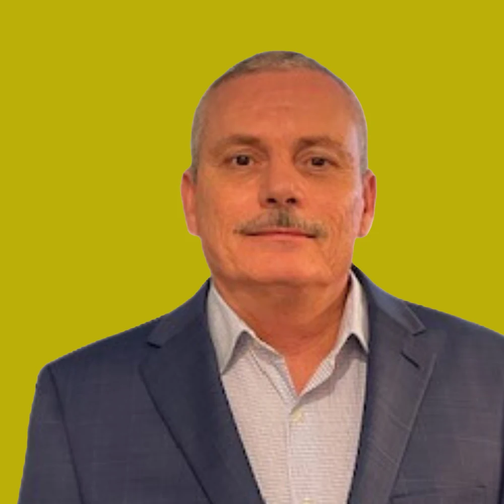 A middle-aged man with short hair and a mustache wearing a blue suit jacket and light shirt stands against a solid yellow background.