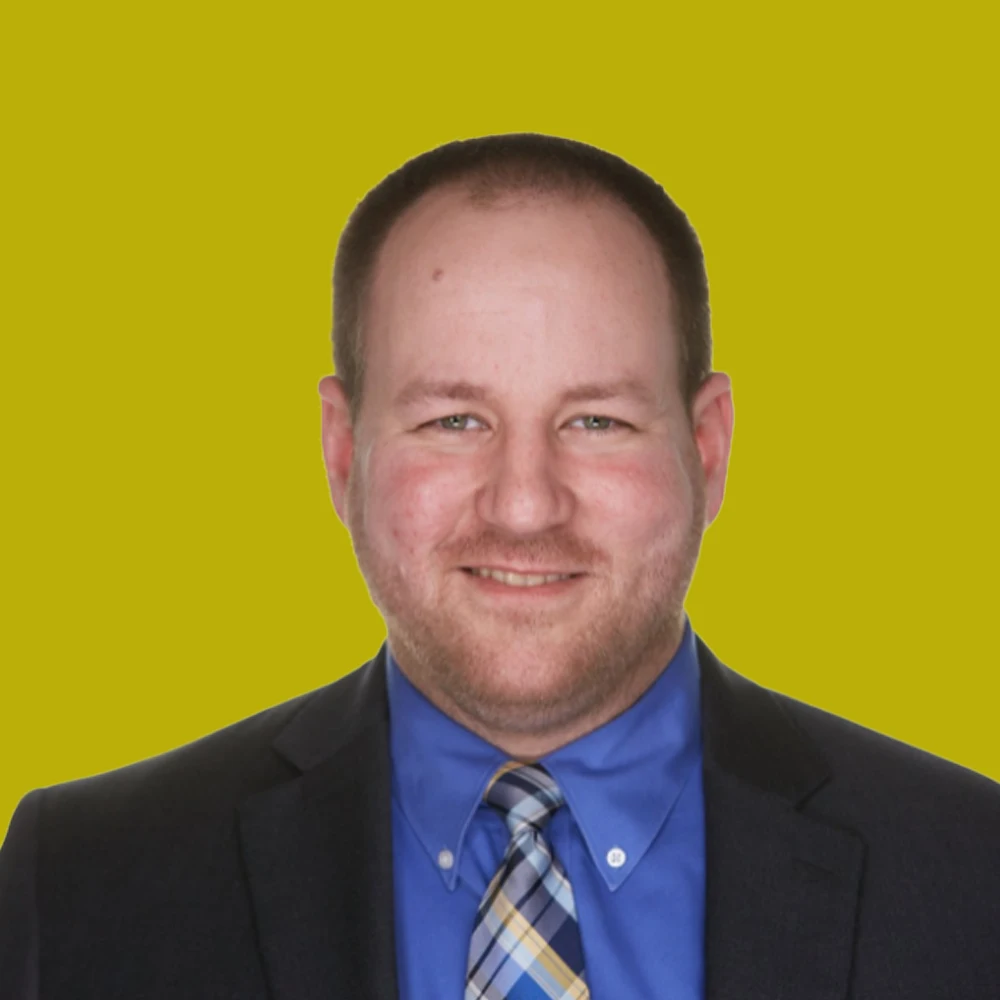 Man in a suit and tie smiles at the camera against a plain yellow background.