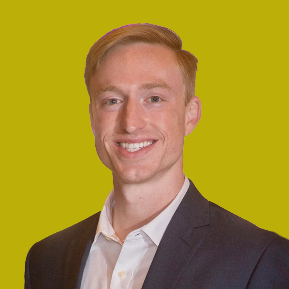 A man with short blond hair in a dark suit jacket and white shirt smiles at the camera against a solid yellow background.