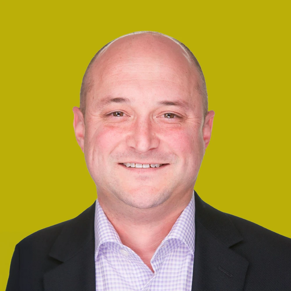 A smiling bald man in a suit jacket and checkered shirt poses against a solid yellow background.