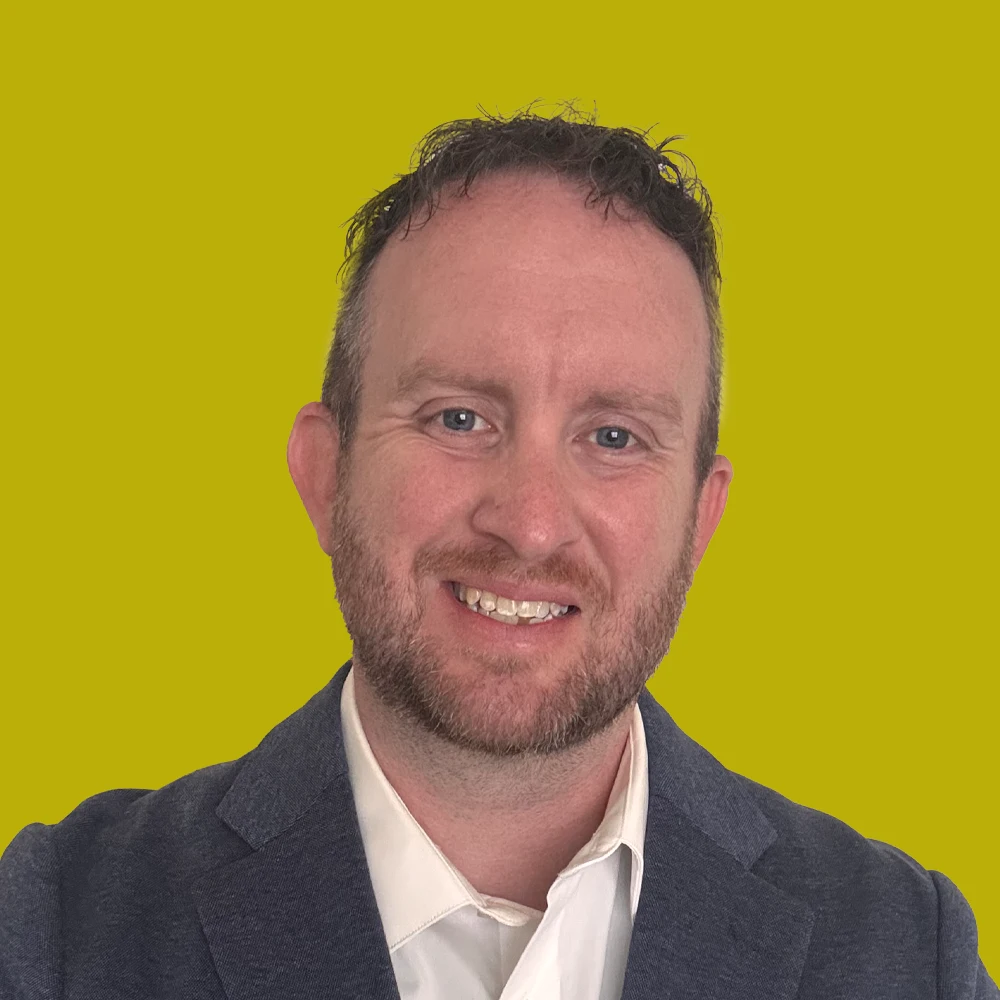 Man with short curly hair and beard, wearing a white shirt and gray blazer, smiling in front of a solid yellow background.