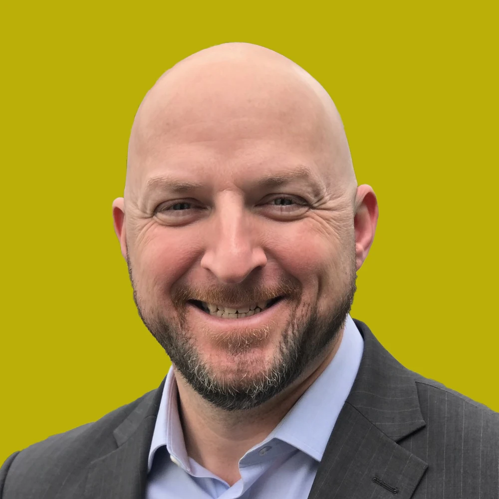 Bald man with a trimmed beard smiling, wearing a suit jacket and light blue shirt, against a solid yellow-green background.