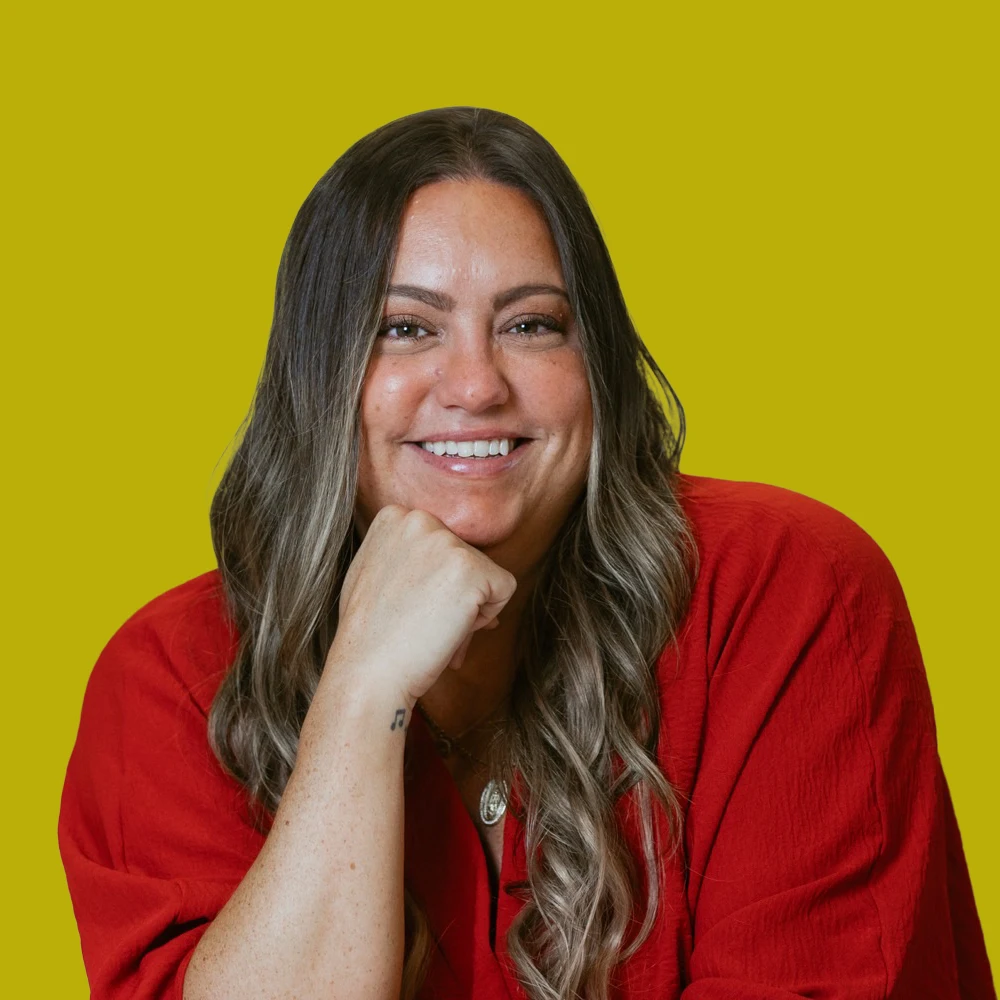 Woman with long brown hair wearing a red top, smiling and resting her chin on her hand, posed against a solid yellow background.