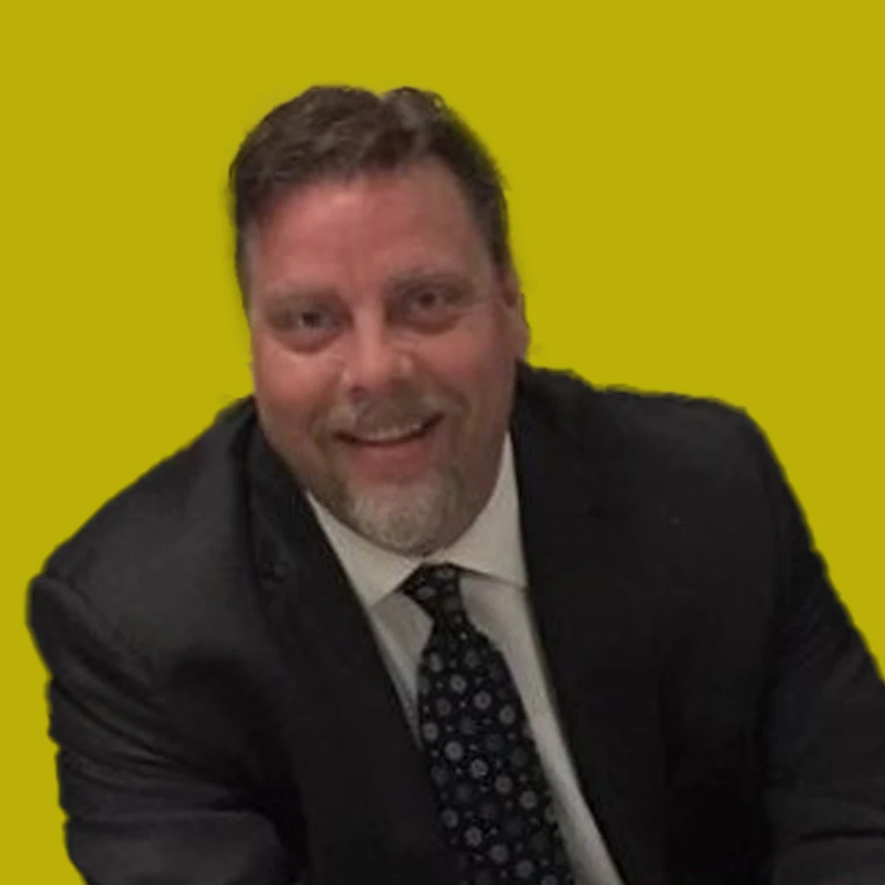 Man in a dark suit and patterned tie smiling, seated against a solid yellow background.