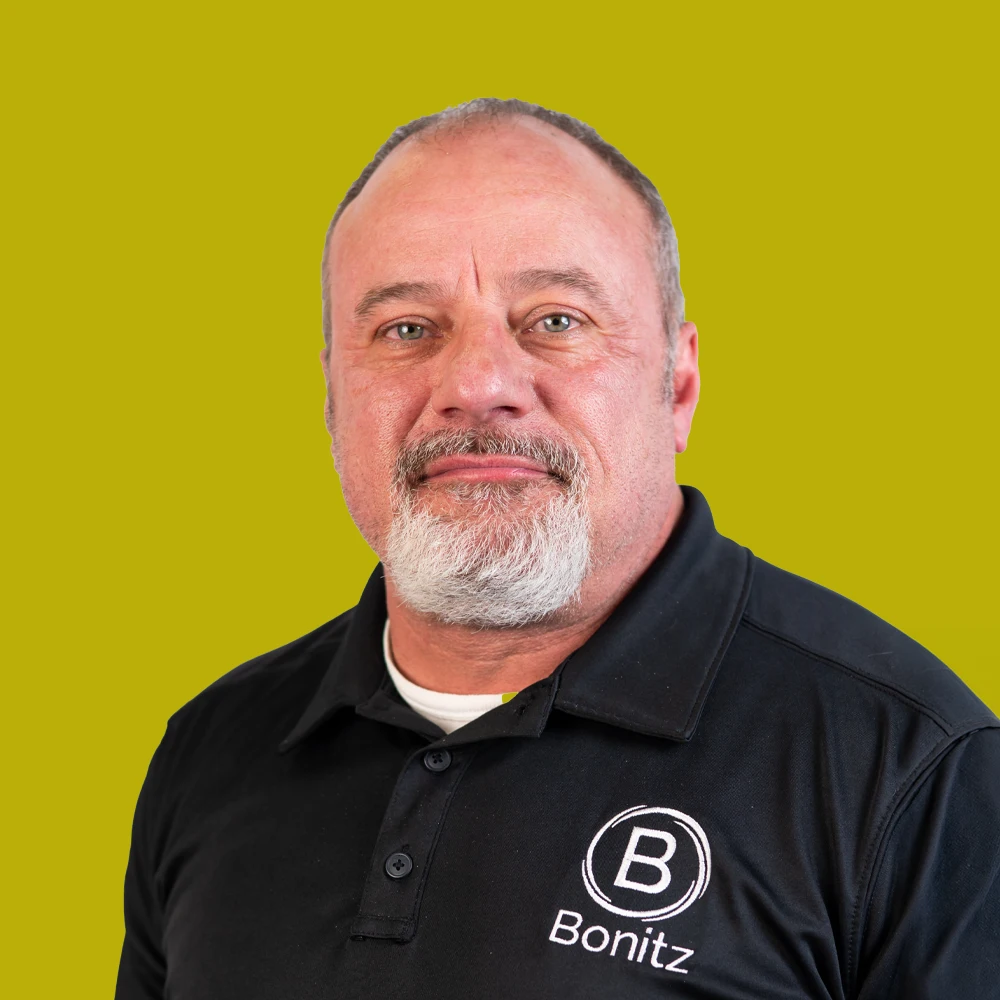 Middle-aged man with short hair and a gray beard wearing a black Bonitz polo shirt, posed against a solid yellow-green background.