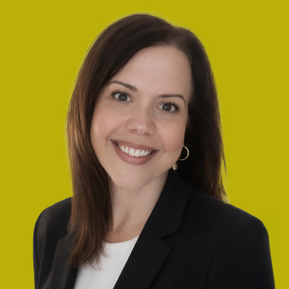 Woman with straight brown hair wearing a black blazer and white top, smiling at the camera, posed in front of a solid yellow-green background.