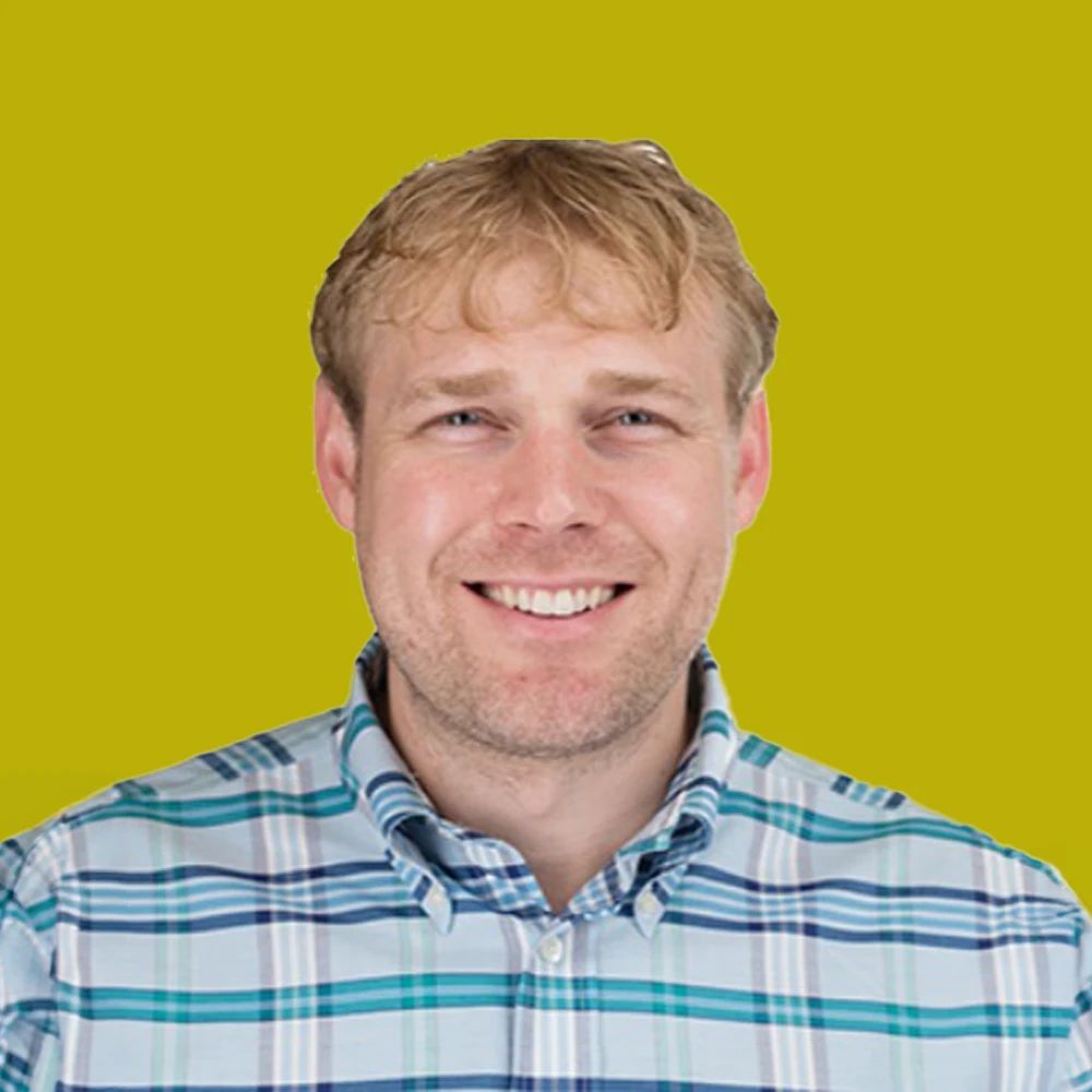 A man with light brown hair wearing a blue and white plaid shirt is smiling in front of a solid yellow background.