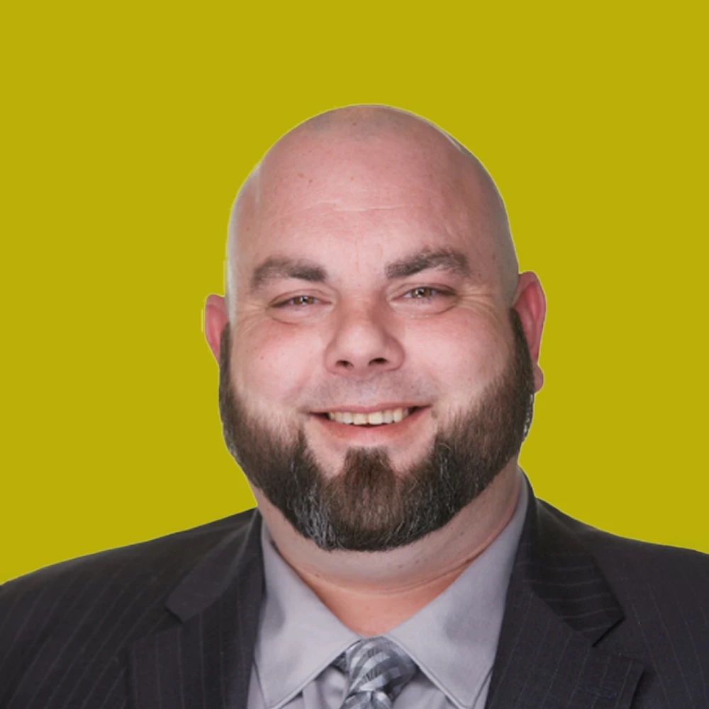 Bald man with a beard in a suit and tie, smiling, in front of a plain yellow background.