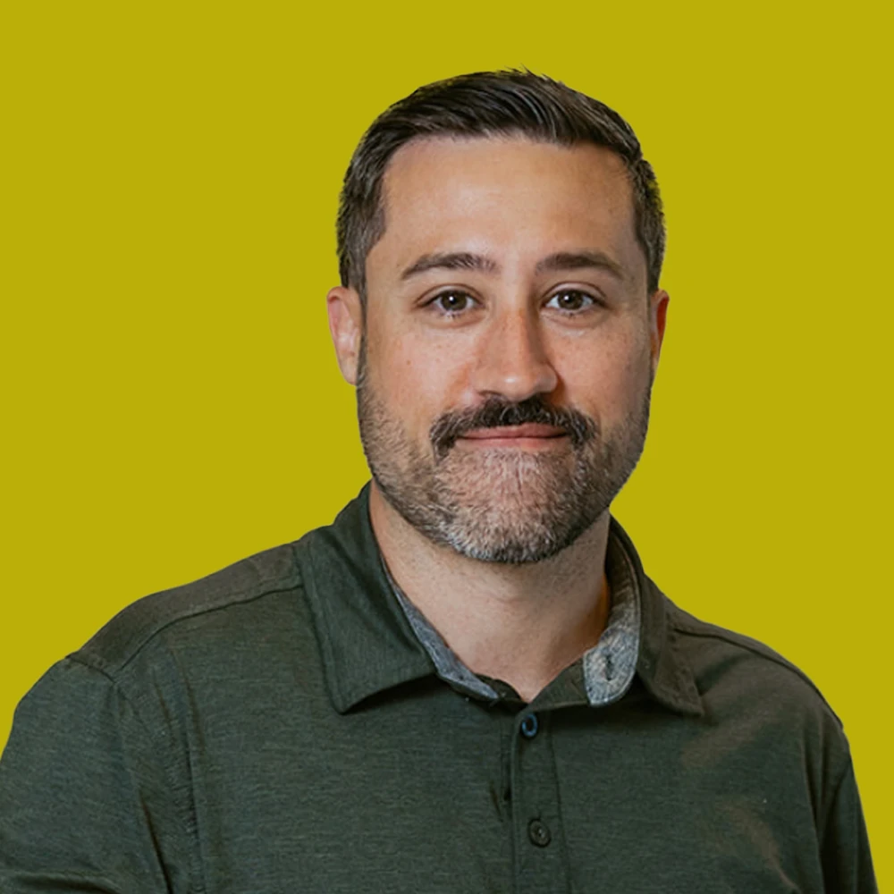 Man with short dark hair and beard wearing a green collared shirt, facing the camera, against a solid yellow background.