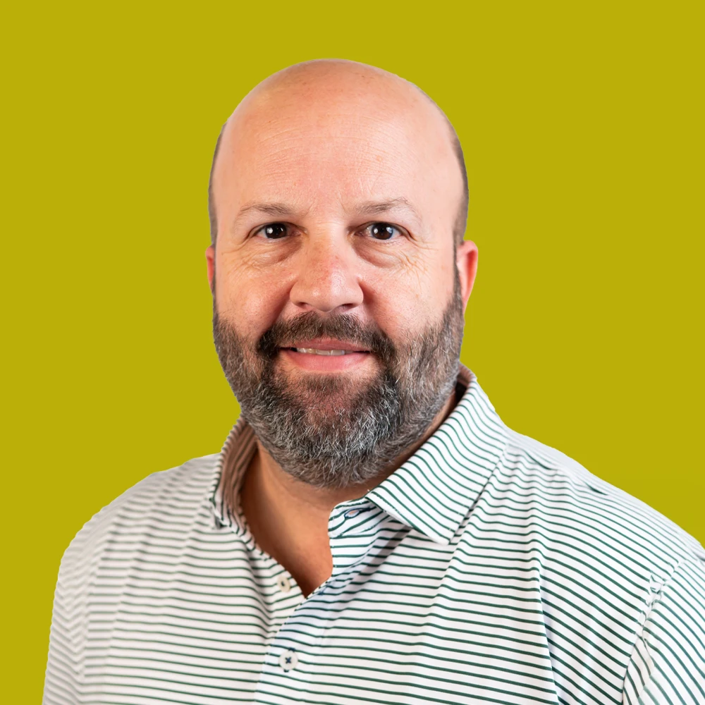 Bald man with a beard wearing a white and blue striped collared shirt, standing in front of a solid yellow-green background.