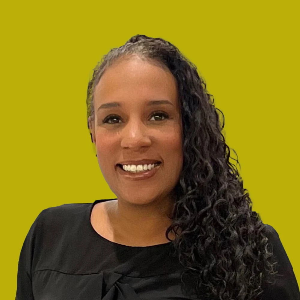 Smiling woman with long curly hair, wearing a black top, posed in front of a solid yellow background.