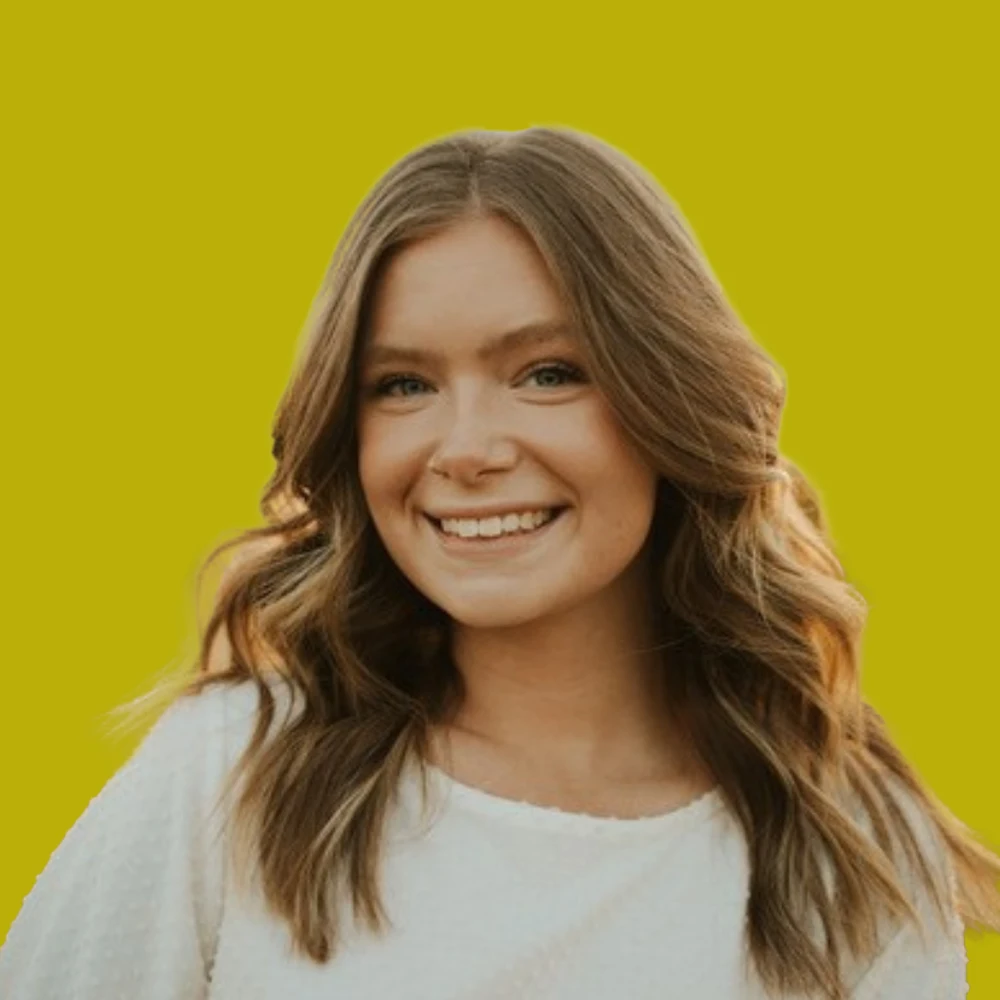 Young woman with wavy brown hair and a white top smiles in front of a solid yellow background.